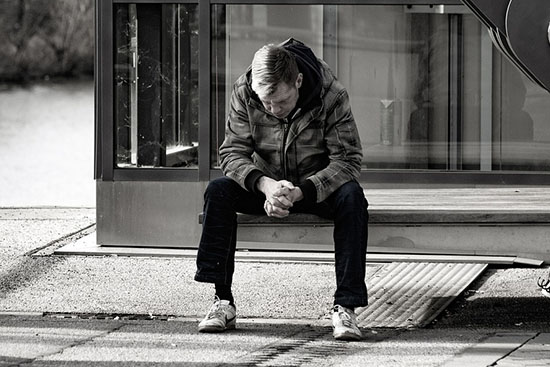 a middle-aged man looks at the ground while sitting on a bench, waiting for the bus