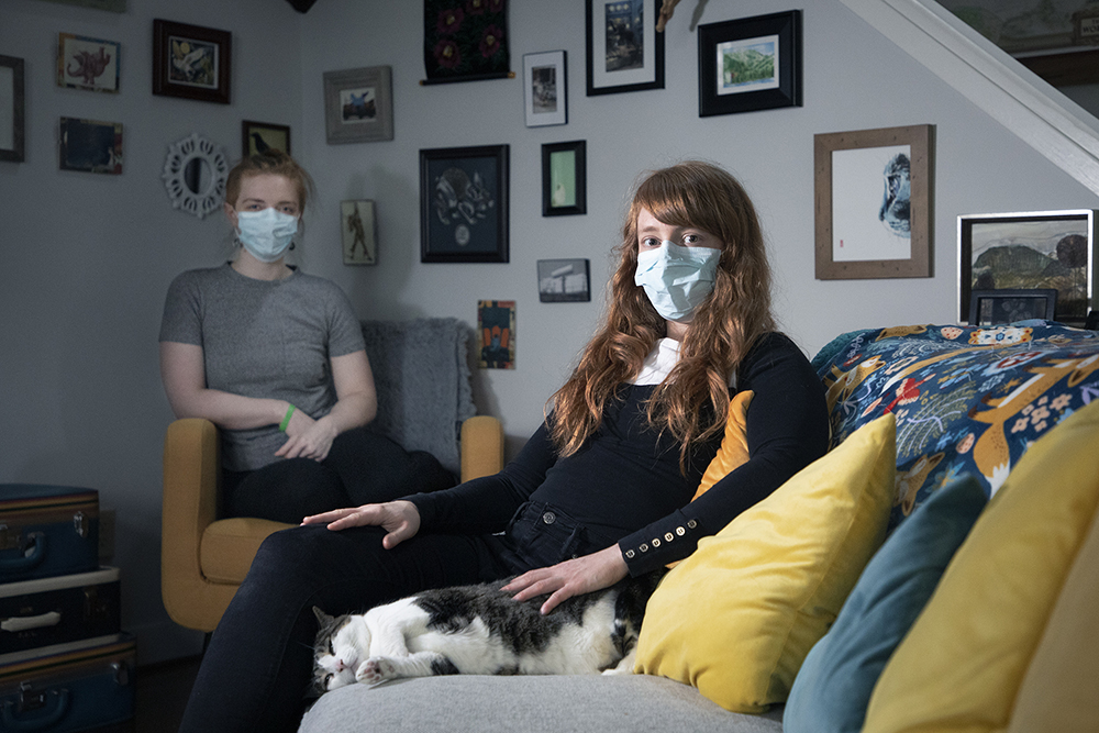 a young, masked woman sits on a couch with her cat; another masked young woman sits behind her in the background