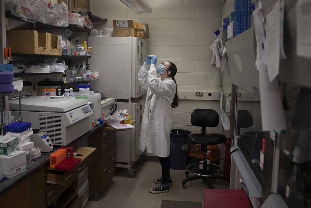 a masked young woman holds up a specimen in a small lab space