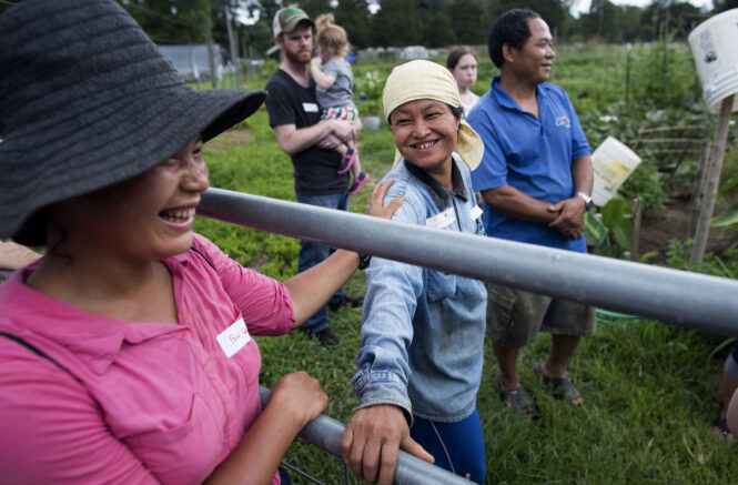 two Karen women stand on each side of a fence; both laugh and one puts her hand on the other's shoulder