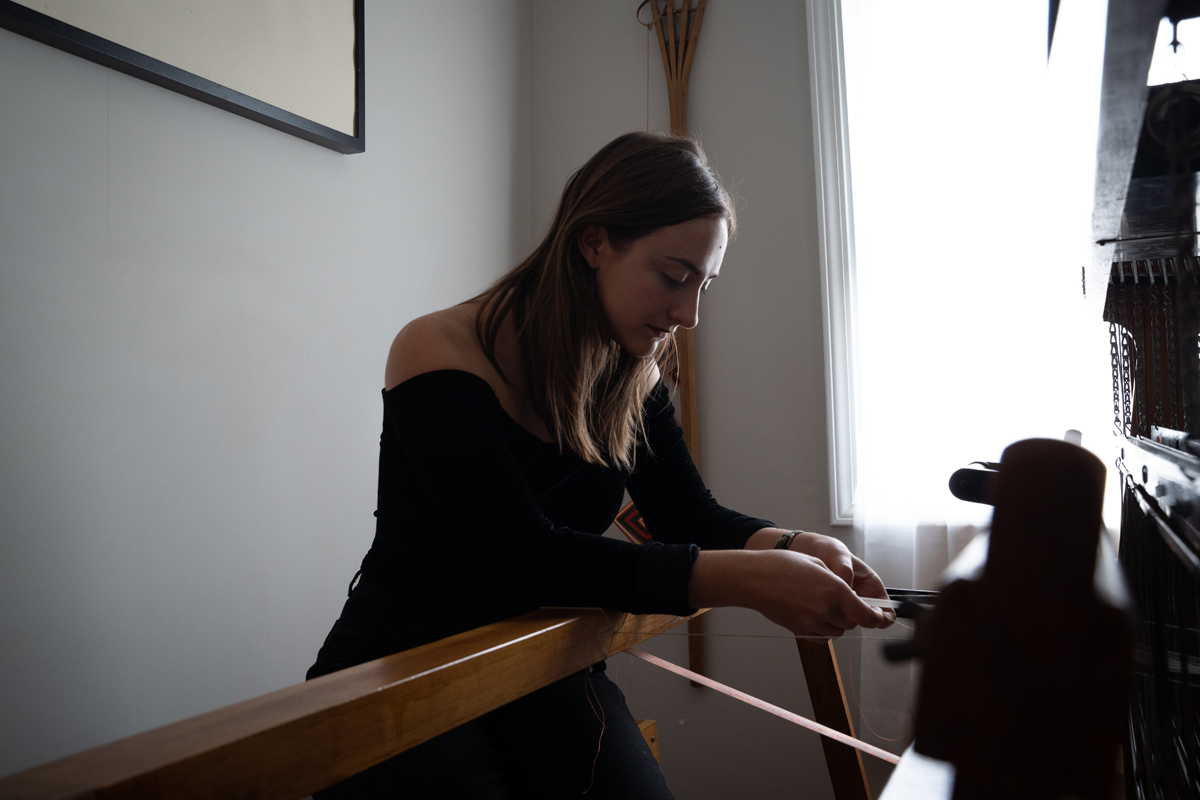 Photo: A young woman in black leans into a large device of wood and metal. She is lit from behind by an open window.