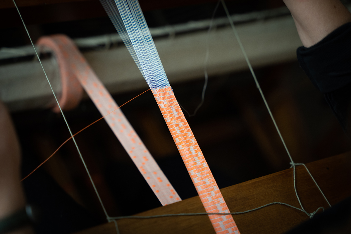 Photo: A long piece of fabric made of interwoven white and orange threads is stretched on a loom. The threads form a pattern reminiscent of a genetic sample test.