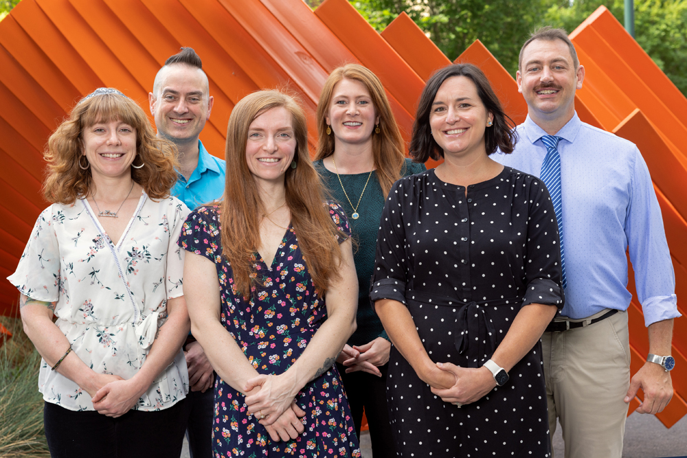 The full-time staff of Endeavors stand in front of an abstract orange sculpture. Left to right (front): Alyssa LaFaro, Corina Prassos, Layla Dowdy; left to right (rear): Darren Abrecht, Carleigh Gabryel, Andrew Russell.