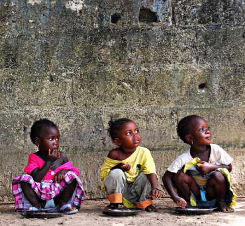 Photo of three children crouched down, eating food off of plates on the ground.