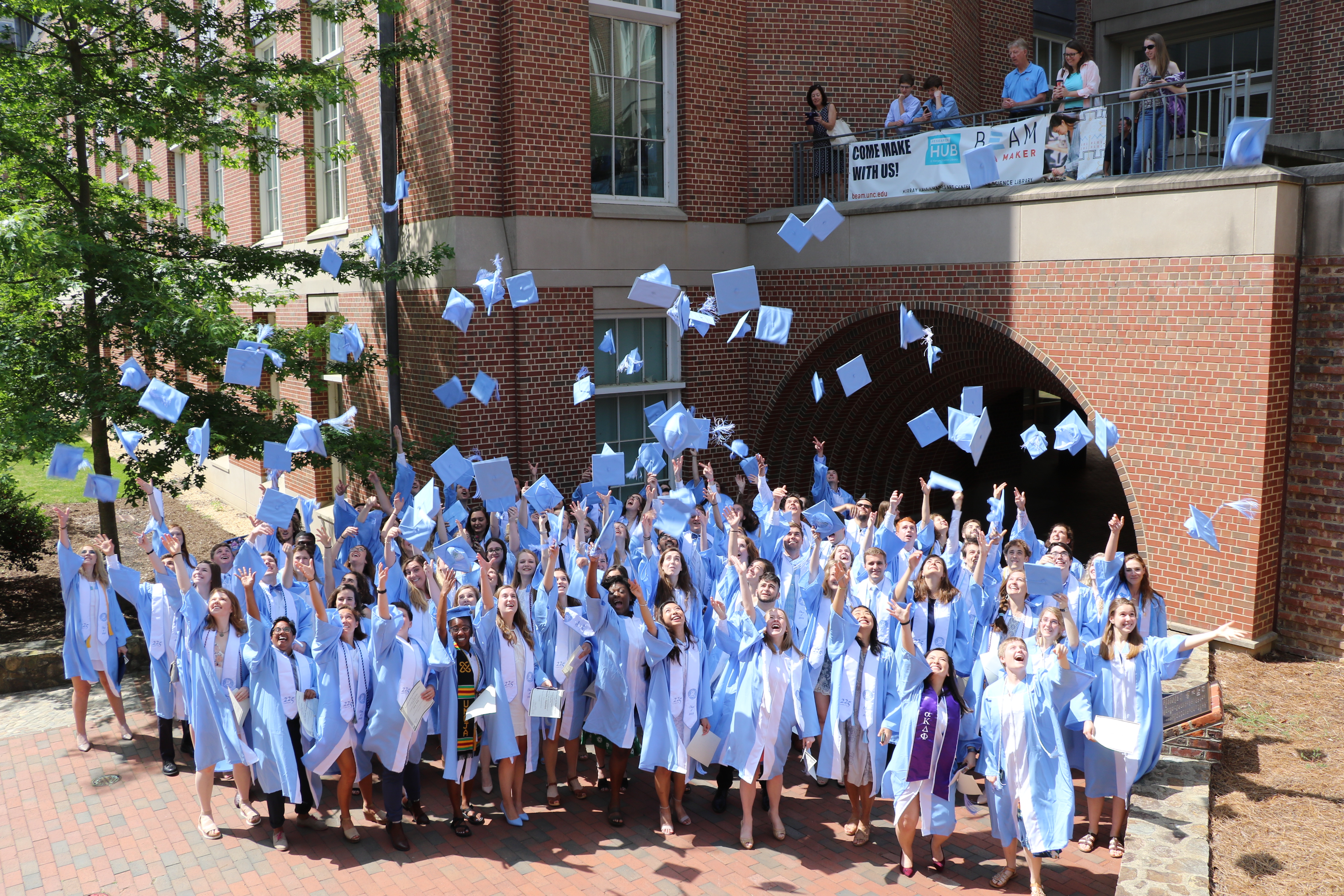 a group of students in Carolina blue caps and gowns throw their hats up in the air in front of Murray Hall on UNC's campus