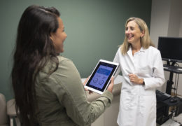Deb Tate (right) speaks with a young woman holding an iPad with the mPWR app on it in an exam room