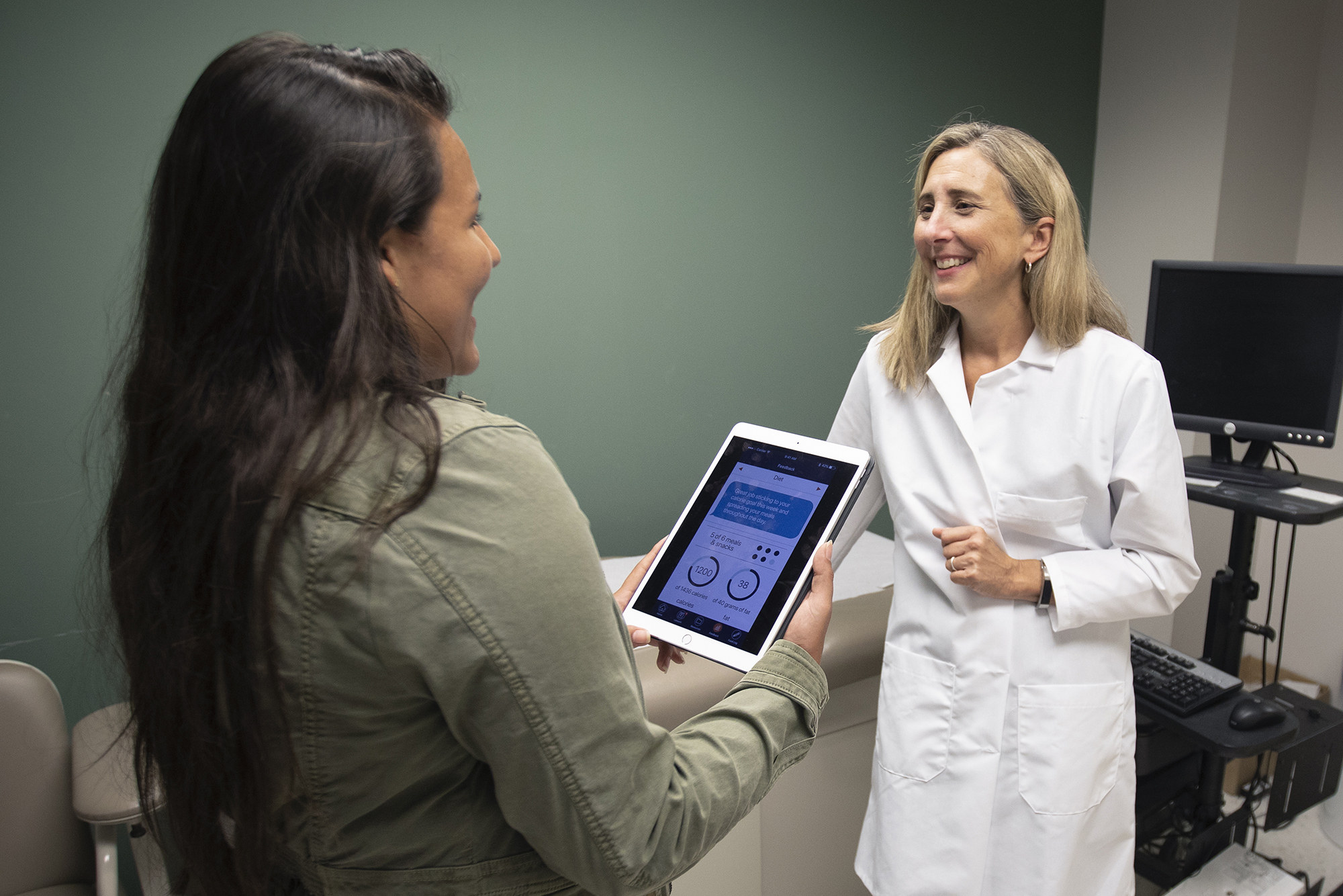 Deb Tate (right) speaks with a young woman holding an iPad with the mPWR app on it in an exam room