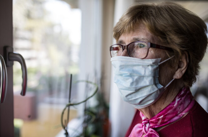 an elderly woman wearing a mask looks out the window