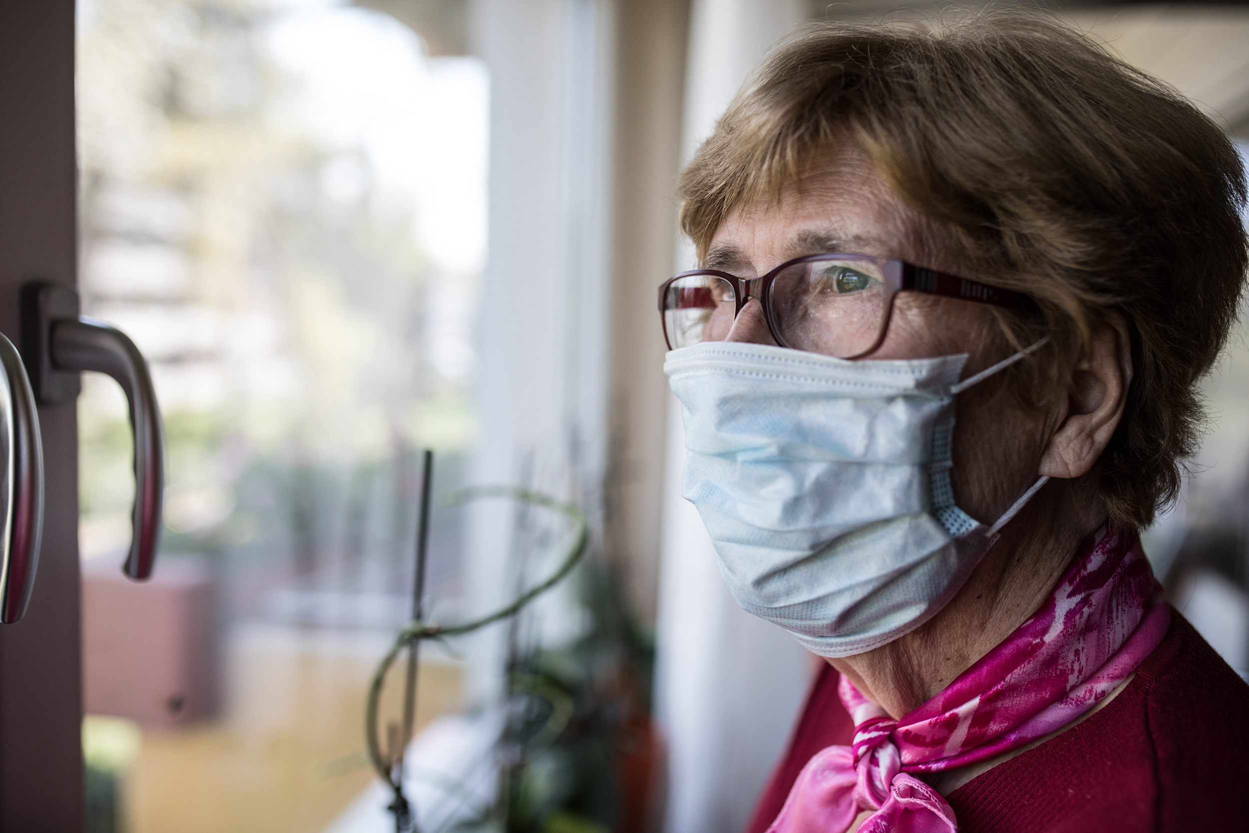 an elderly woman wearing a mask looks out the window