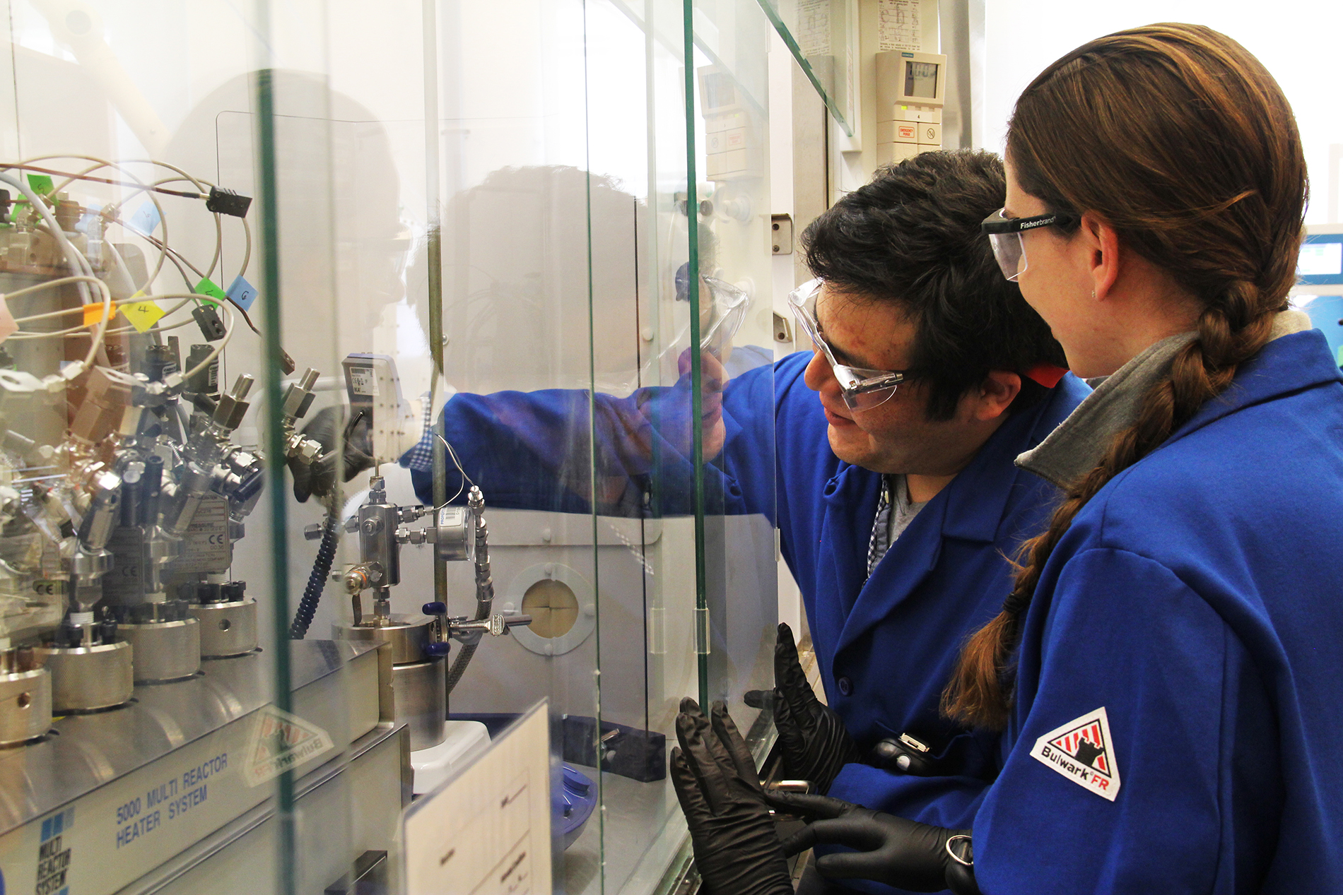 a woman and a man in blue lab coats lower the pressure gage on technical equipment inside a lab