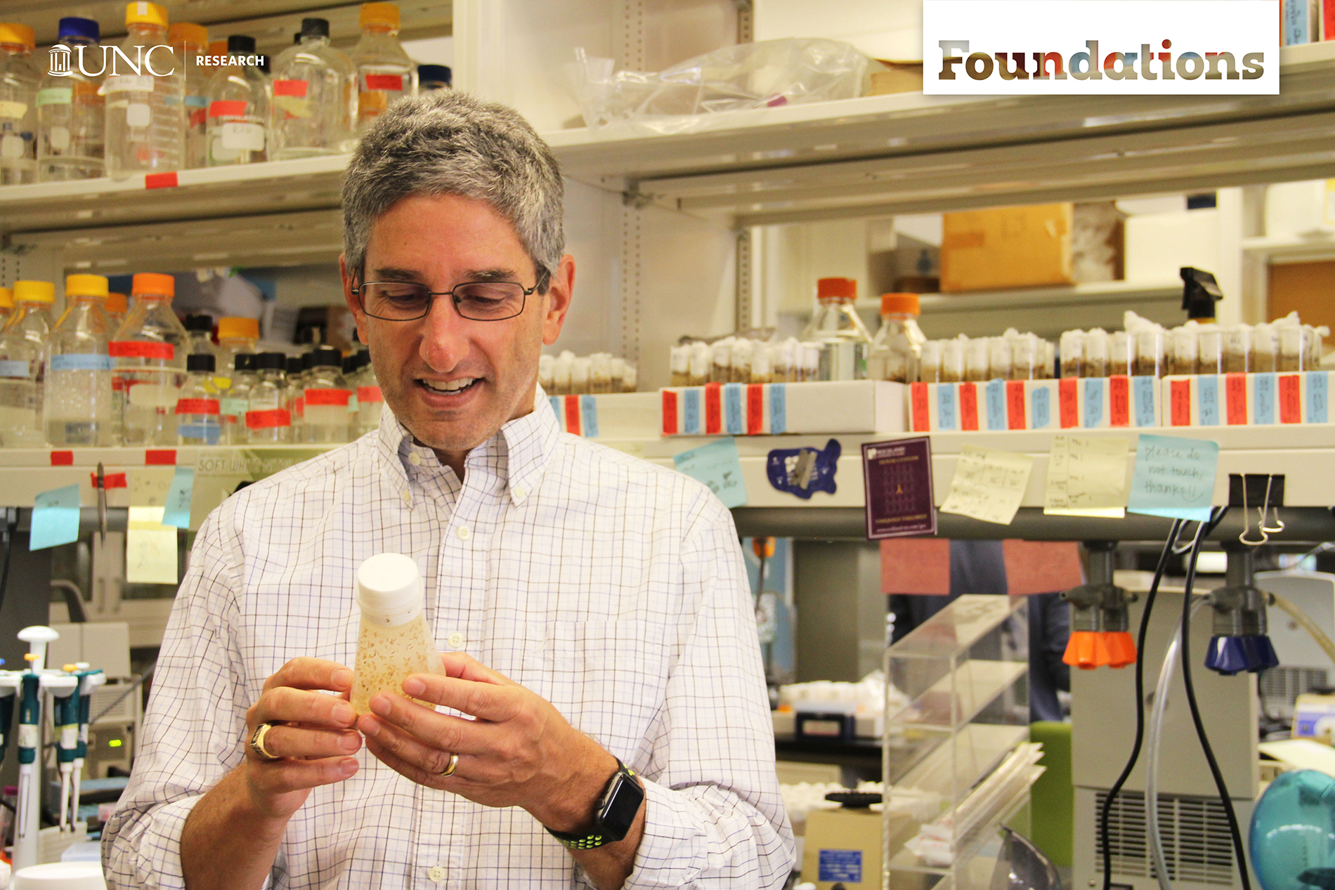 a 40-something male holds a container of fruit flies -- shelves with colorful labels are in the background