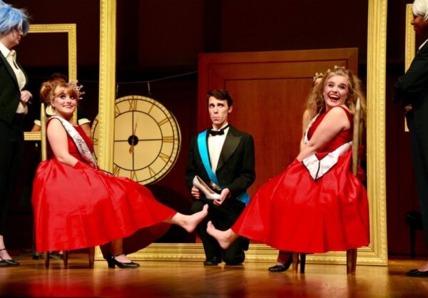 a young man in a tuxedo holds a show as two young women in bright red dresses sit with their feet pointed out on either side of him