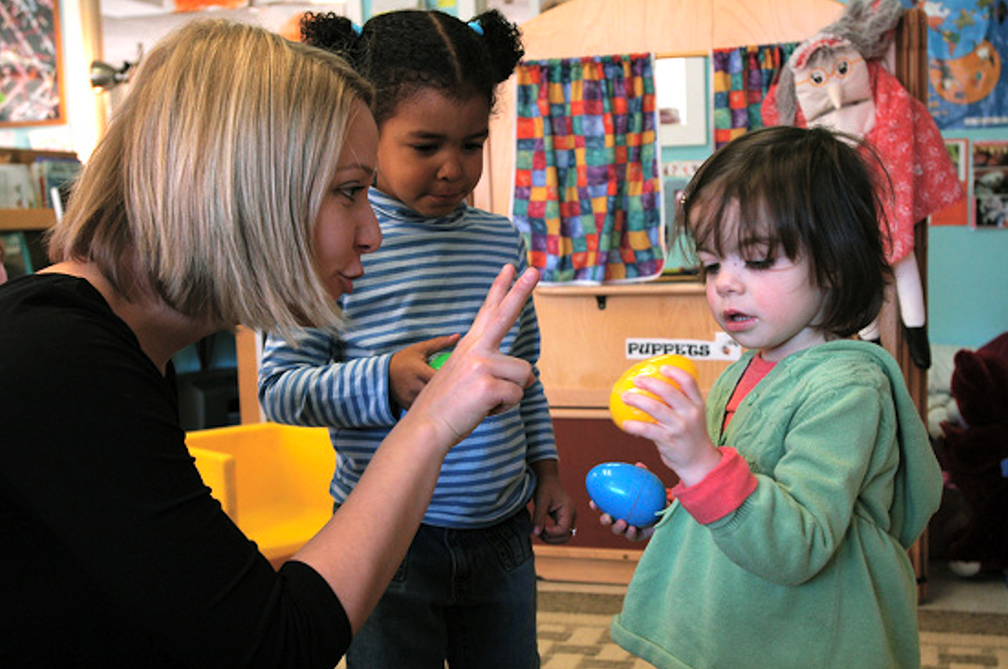 a blond woman holds two fingers up to a toddler, trying to teach her how to count