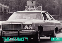 Old image of a researcher sitting in a car, with a sign on the top of it saying "7; Driver Education. Captial Area Multi-Vehicle Laboratory"