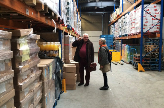a woman talks with a male food bank official as they stand in a warehouse among shelves of canned food