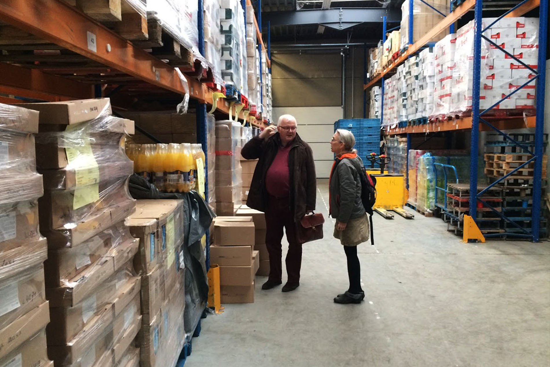 a woman talks with a male food bank official as they stand in a warehouse among shelves of canned food