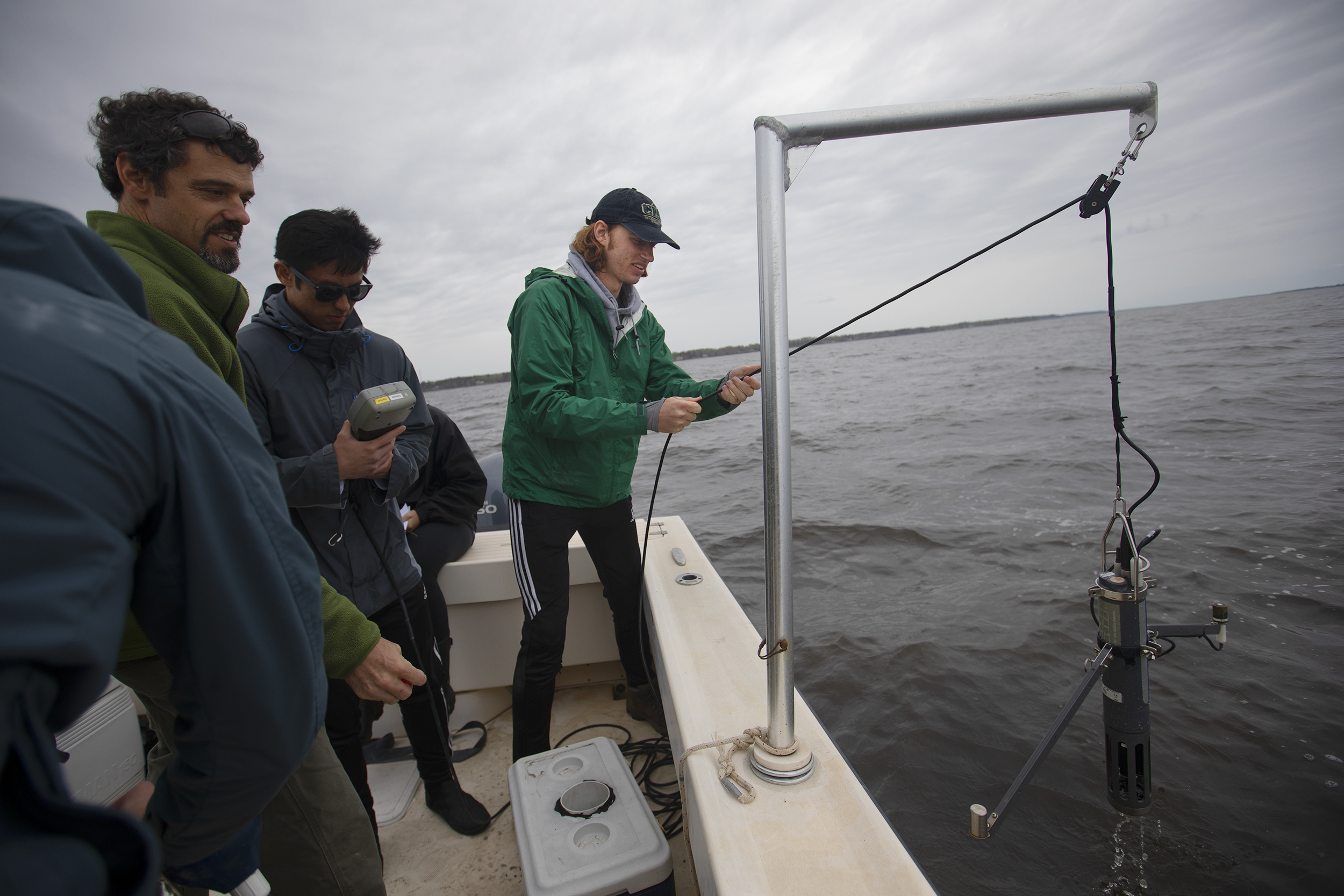 A student stands next to the edge of the boat and uses a pulley system to raise a metal instrument from the estuary water.