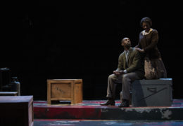 A male and female actor sit on military style crates while talking on stage..
