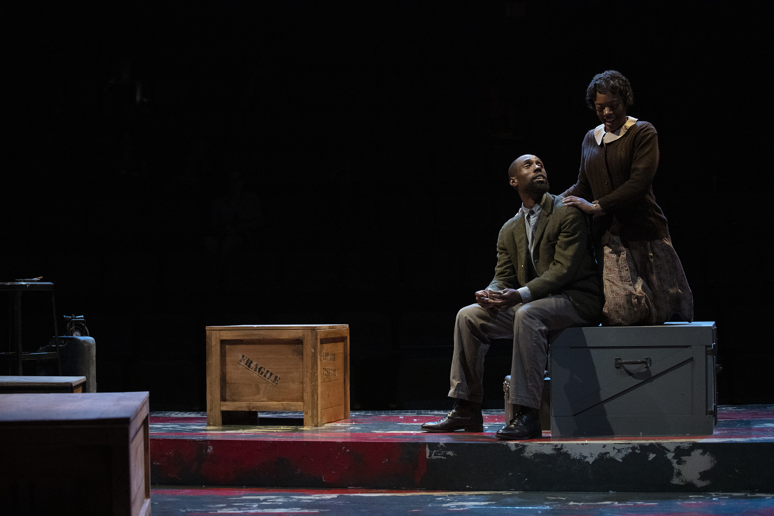 A male and female actor sit on military style crates while talking on stage..