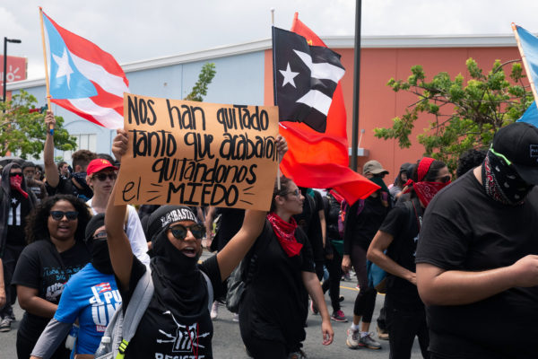a young Puerto Rican woman marches in a protest with a sign that says, "Nos han quitado tanto que arabaton quitandonos el miedo"