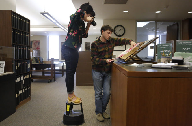 Sarah stands on a library stool while taking a photo of hospital ledgers, held by Lucas in the North Carolina State Archives.