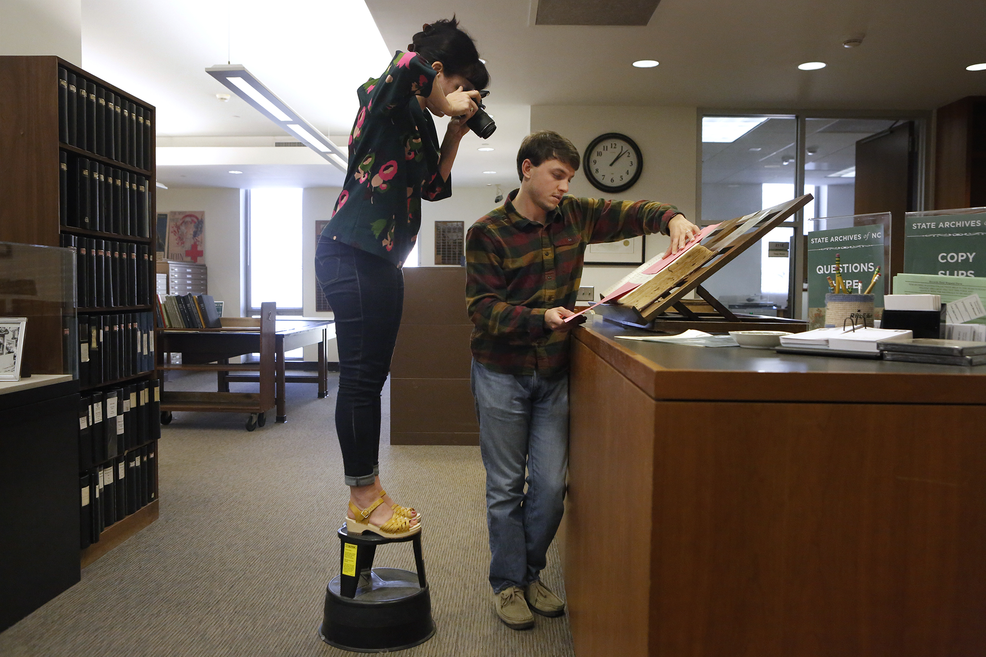 Sarah stands on a library stool while taking a photo of hospital ledgers, held by Lucas in the North Carolina State Archives.