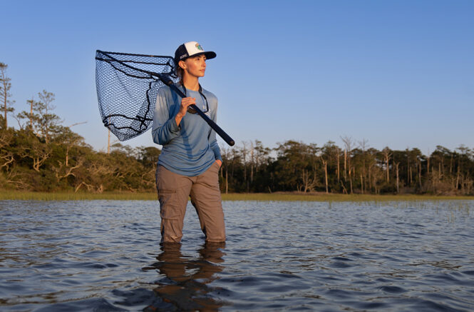young woman holding a fishing net over her shoulder looks out at the water