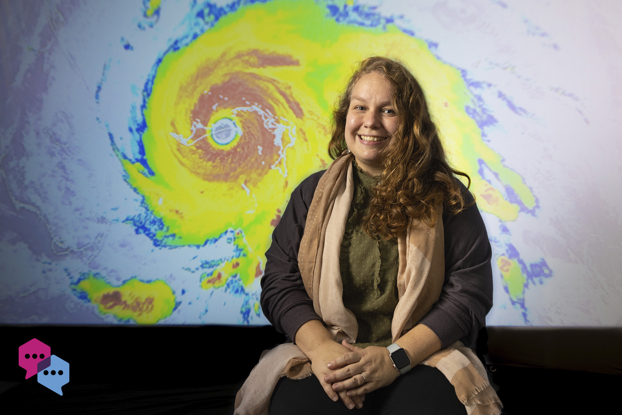 Caela O'Connell sits in front of a projection of a hurricane