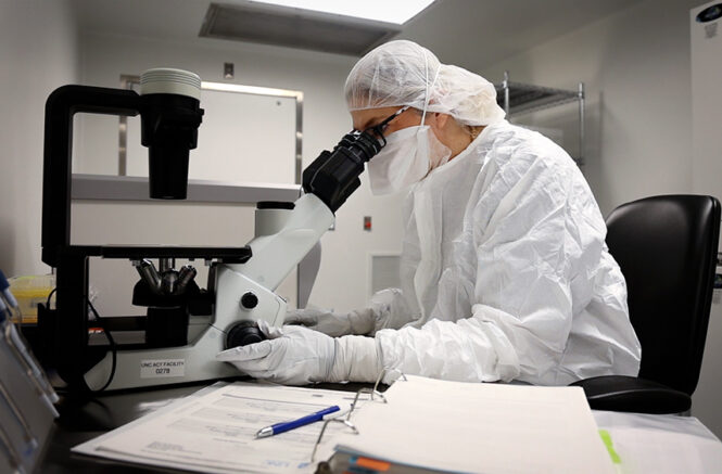 a researcher in head-to-toe white PPE looks through a microscope
