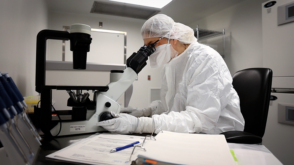 a researcher in head-to-toe white PPE looks through a microscope