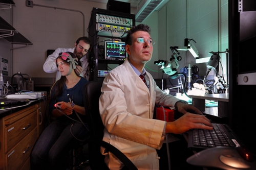 Photo of Flavio Frohlich looking at him computer as graduate student Michael Boyle attaches a head piece to graduate student Kristin Sellers.