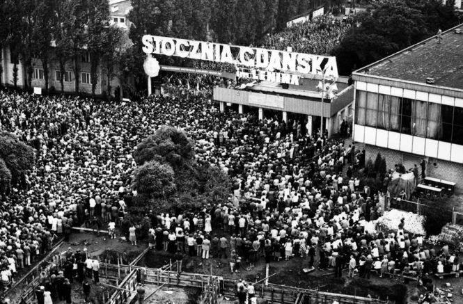 A large crowd of people gather in front of a sign that reads "Stoczni Gdańskiej im. Lenina."