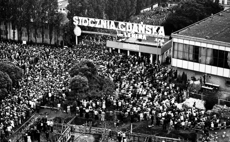 A large crowd of people gather in front of a sign that reads "Stoczni Gdańskiej im. Lenina."