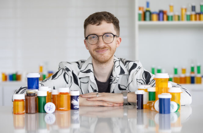 a young man leaning against a white table covered in colorful prescription bottles