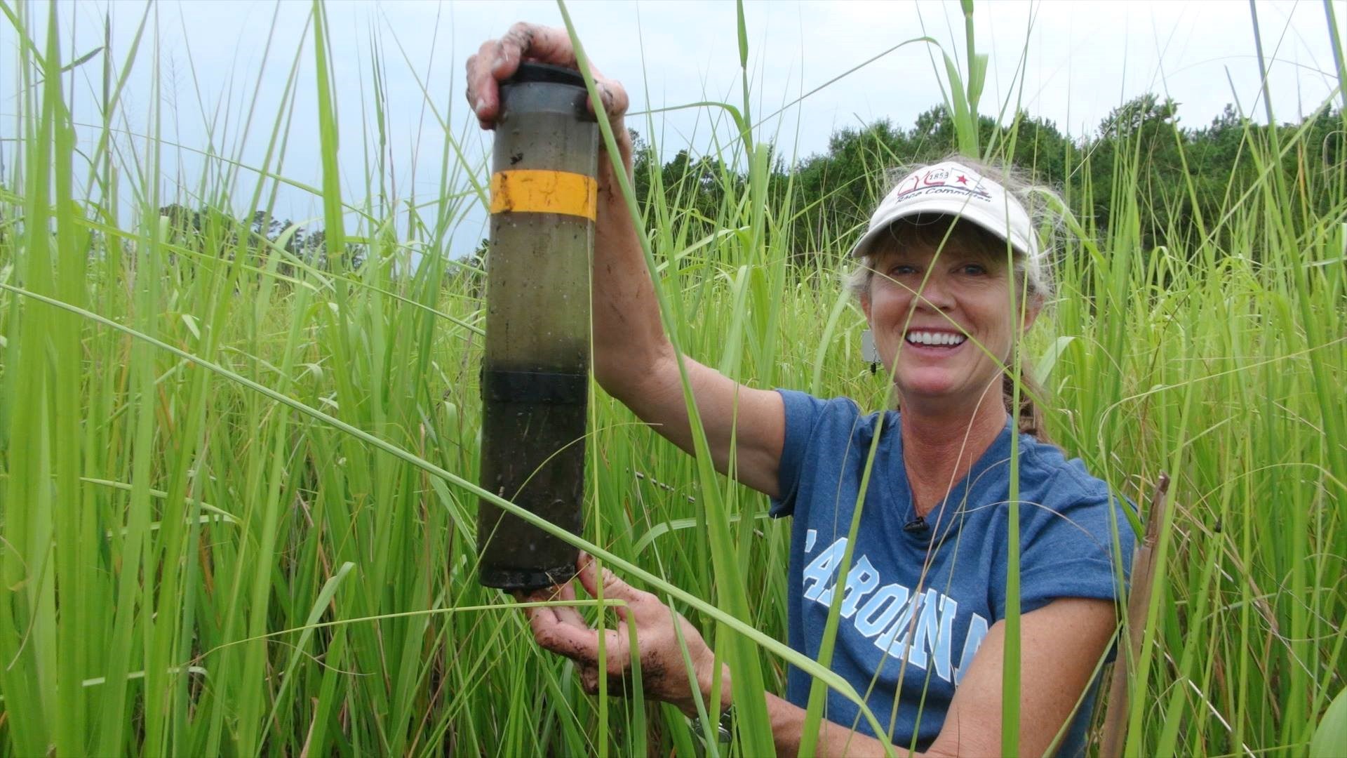 a woman stands in a marsh and holds up a big piece of sediment