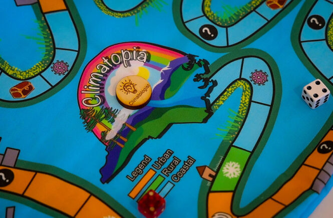 Photo of the center of the fabric Climatopia game board with a wooden game piece. The fabric game board depicts an outline of the state of North Carolina on a blue background with a rainbow arching over the outline.