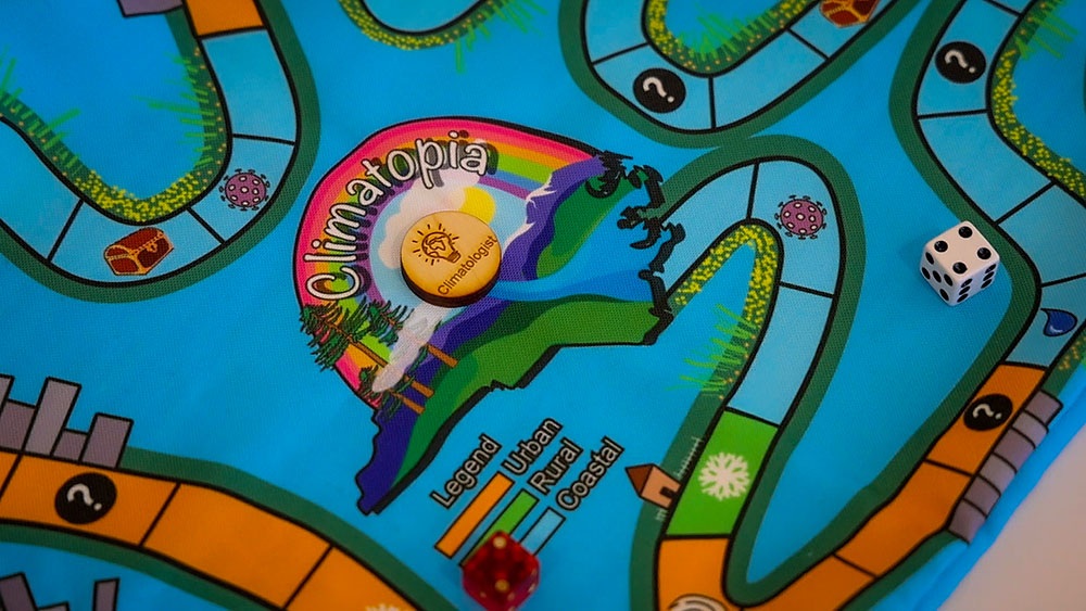 Photo of the center of the fabric Climatopia game board with a wooden game piece. The fabric game board depicts an outline of the state of North Carolina on a blue background with a rainbow arching over the outline.