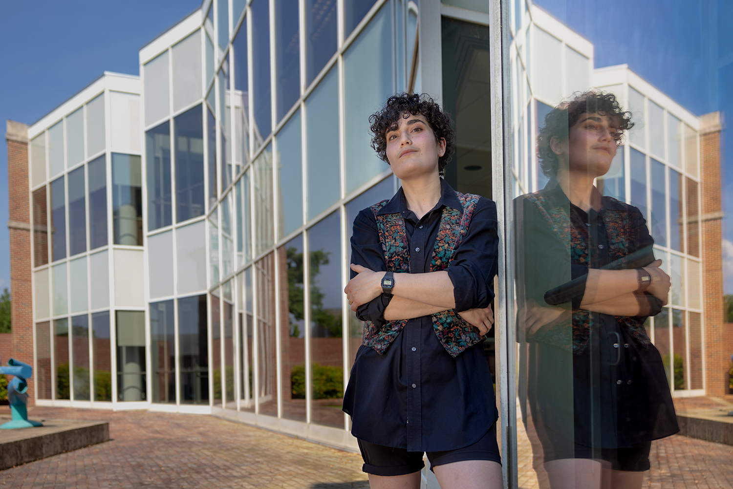 Portrait of Suad Jabr. They are outside, leaning against a reflective surface with their arms crossed.