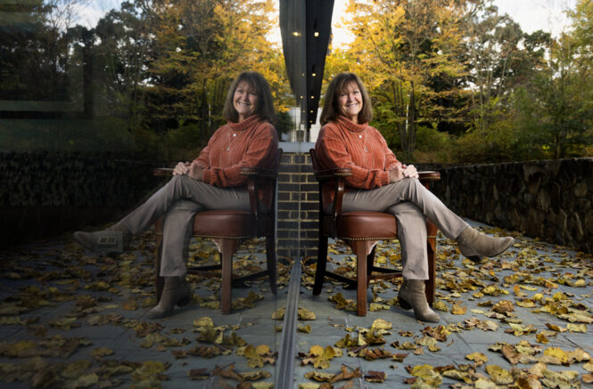a woman sits in a chair surrounded by leaves, her reflection in a nearby window
