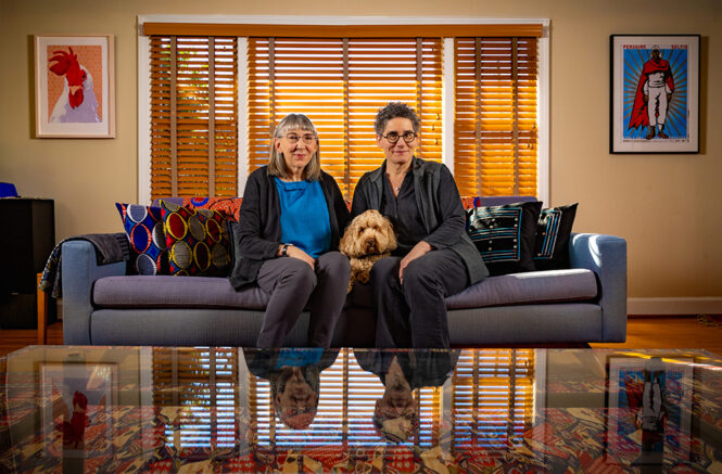 Florence Babb and Victoria Rovine are smiling while sitting on a sofa in their living room. Their dog, Harry, is sitting between them.