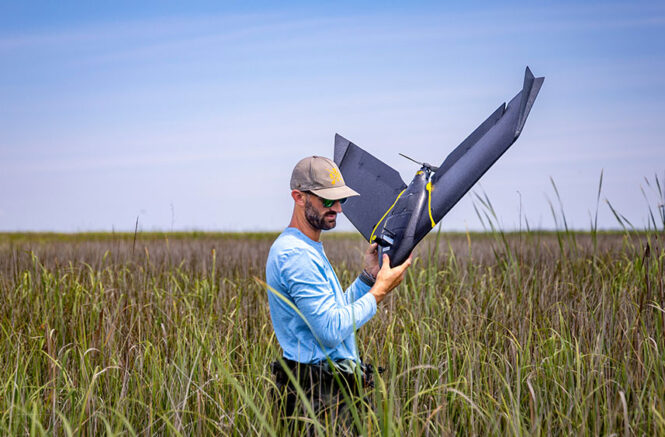 A man carries a drone out of a marsh.