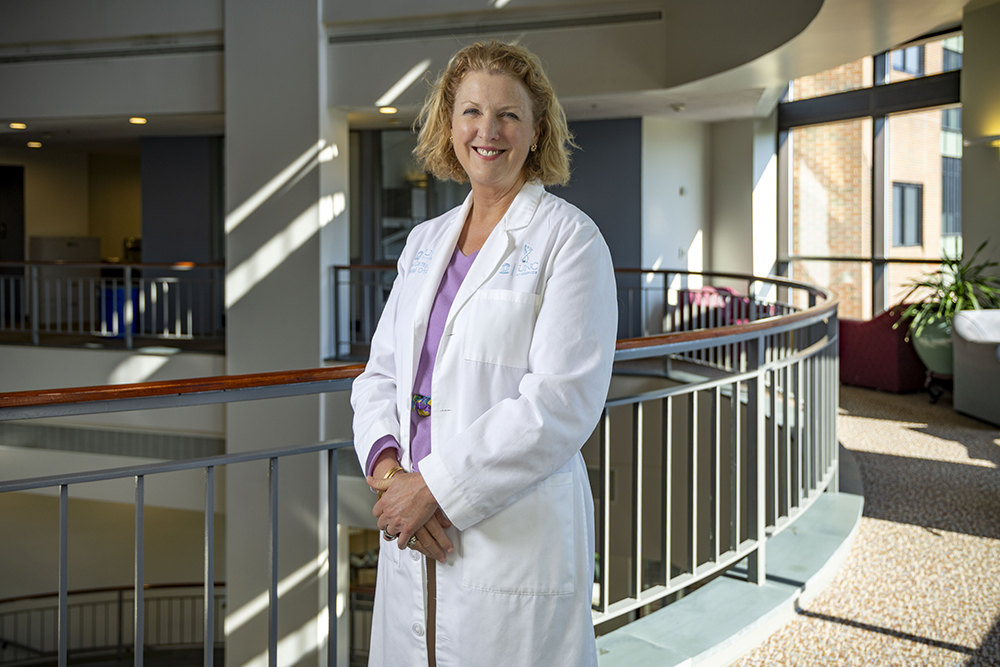 A woman in a white lab coat smiles while standing in a hallway of a medical office building.