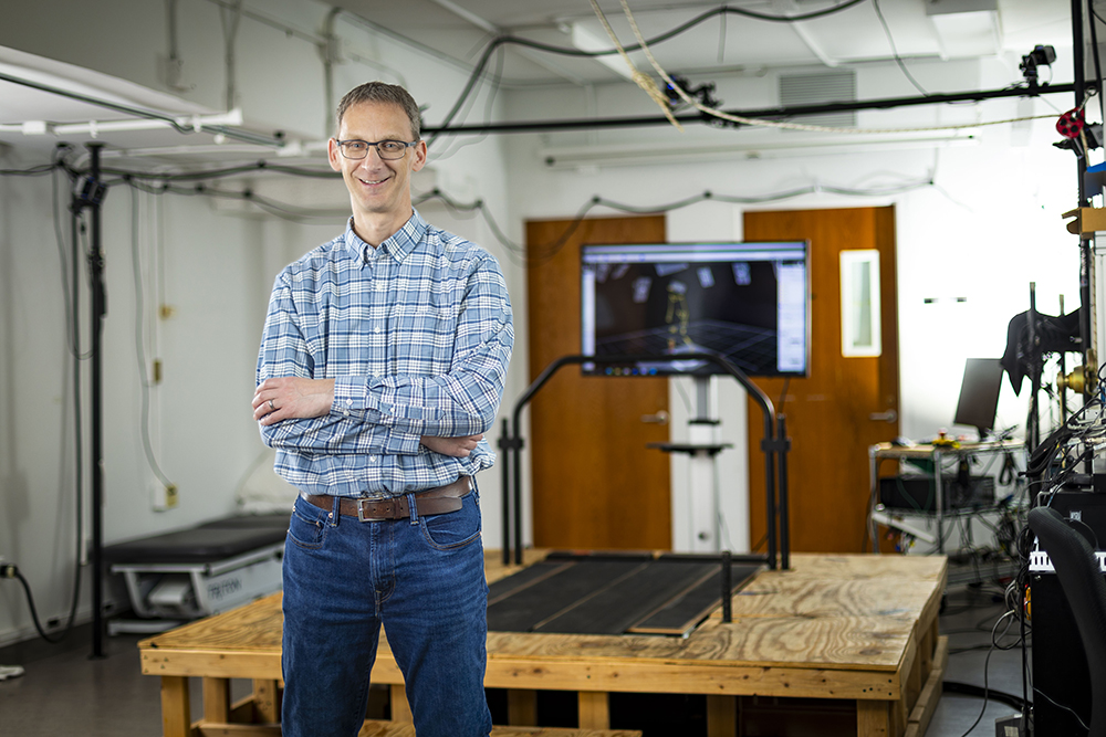 A man smiles with his arms crossed while standing in front of a tread mill in a research lab.
