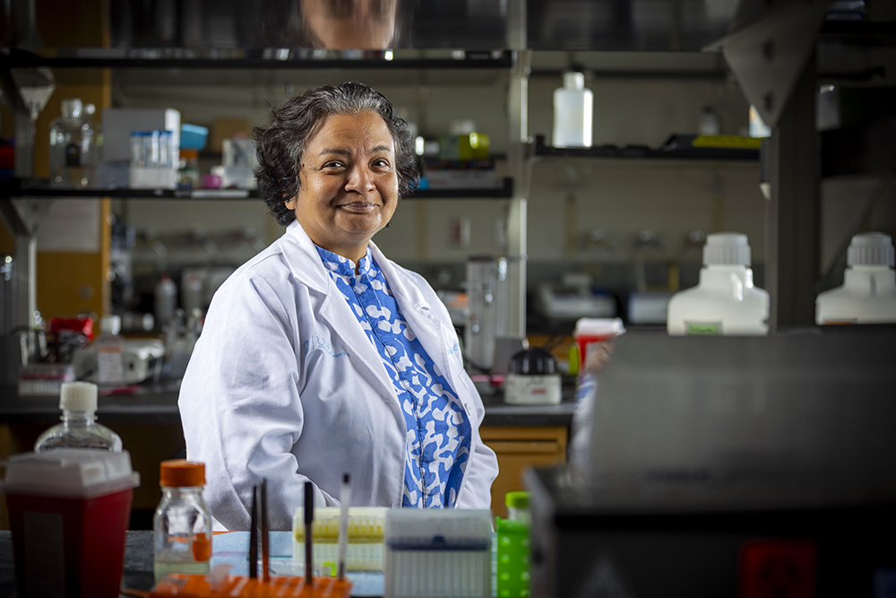 A woman in a white lab coat smiles while standing at a lab bench.