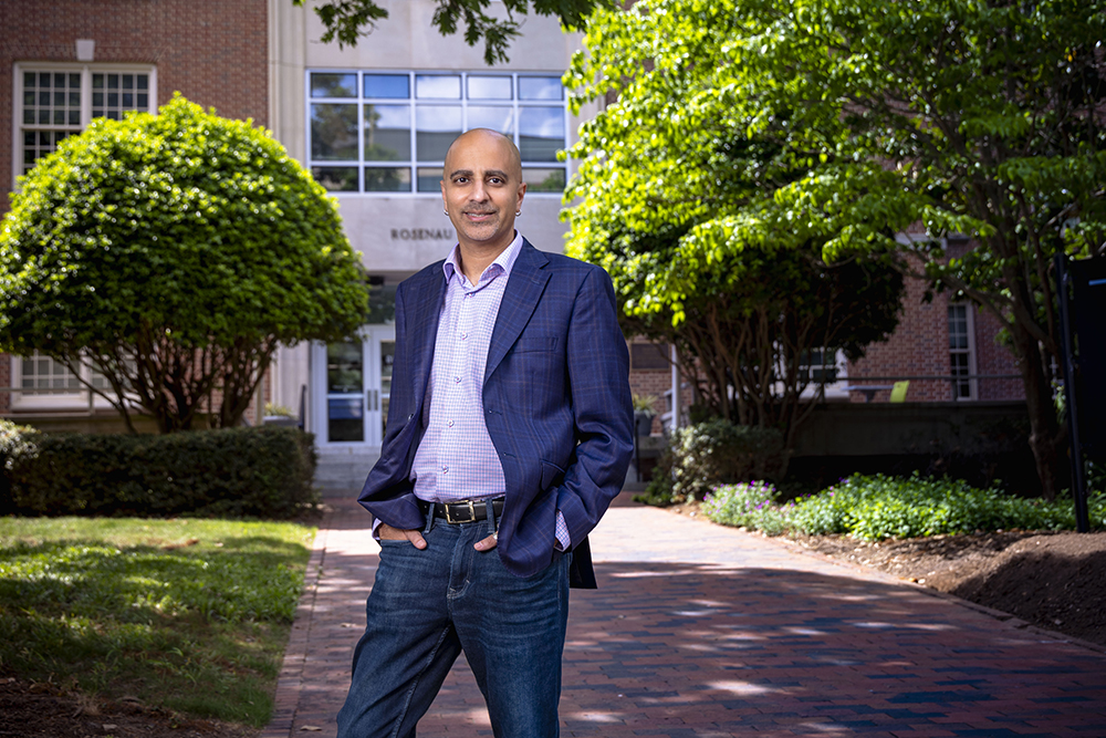 A man with his hands in his pocket smiles at the camera while standing in front of a brick building on a university campus.