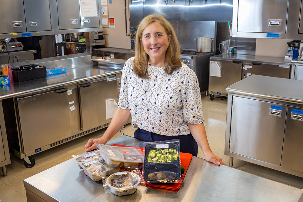 A women stands in a kitchen with stainles steel appliances while smiling at the camera.