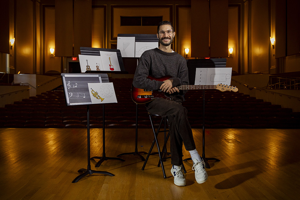 A man sits on stage holding a guitar and surrounded by music stands.