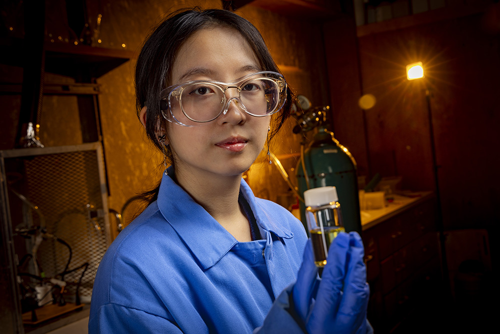 A female chemistry student wearing safety goggles and a blue lab coat holds a glass container.