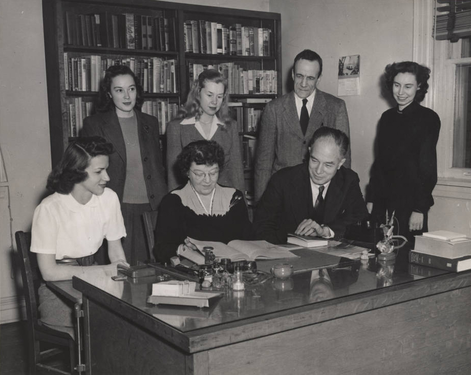 Photo: A woman seated at a desk examines a book. She is surrounded by four women and two men in business attire. Behind them are two large bookshelves.