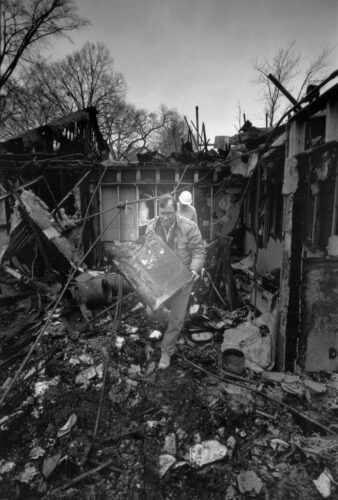 Photo: Two men emerge from a burned-out building. The man in the background is wearing a white hard hat. The man in the foreground is carrying a large rectangular object.